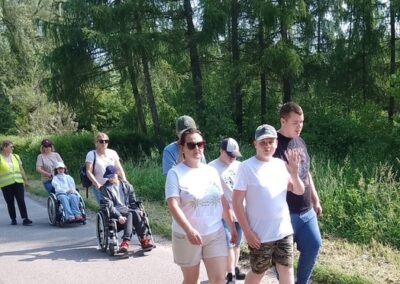 Grupa osób spacerująca po leśnej drodze, w tym dwie osoby na wózkach inwalidzkich.