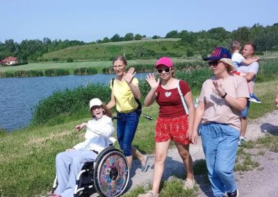 Grupa osób spaceruje nad stawem, jedna osoba na wózku inwalidzkim, zielone wzgórza w tle.