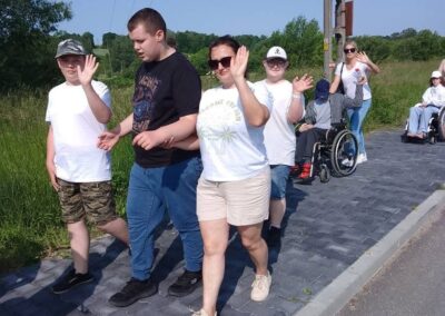 Grupa osób spacerujących po wiejskim chodniku, w tym osoby na wózkach inwalidzkich.