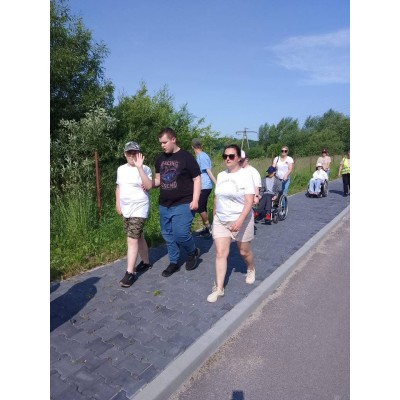 Grupa osób, w tym osoby na wózkach inwalidzkich, spaceruje po chodniku wśród zieleni.