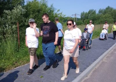 Grupa osób, w tym osoby na wózkach inwalidzkich, spaceruje po chodniku wśród zieleni.