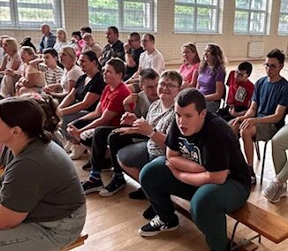 Grupa osób siedzi na ławkach w jasnej sali z dużymi oknami, prawdopodobnie w pomieszczeniu sportowym.