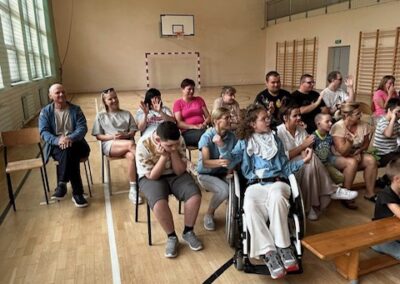 Grupa osób w hali sportowej siedzi na krzesłach i ławkach, w tym osoba na wózku.