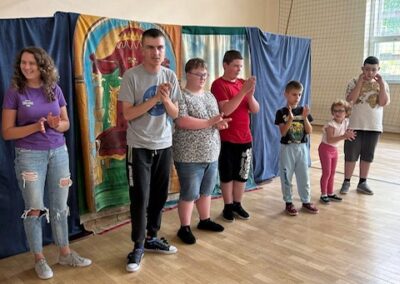 Grupa siedmiu osób stoi i klaszcze w sali gimnastycznej przed kolorową zasłoną.