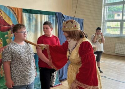 Kobieta w stroju króla dotyka kijem ramienia chłopca w sali gimnastycznej z dwoma innymi chłopcami.
