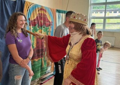 Kobieta w królewskim stroju dotyka berłem bark młodej dziewczyny w hali sportowej.