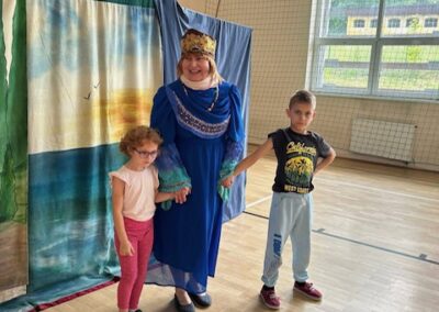 Kobieta w niebieskiej sukni i koronie stoi z dwójką dzieci na sali gimnastycznej przy zasłonie.