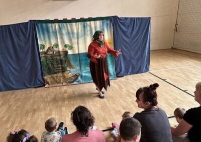 Kobieta występująca na sali gimnastycznej przed dziećmi i dorosłymi na ławkach, za nią dekoracja.