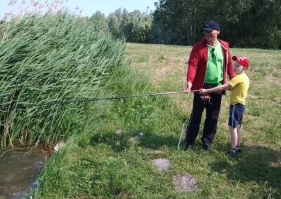 Mężczyzna i chłopiec wędkują nad brzegiem zbiornika otoczonego trawą i trzcinami.