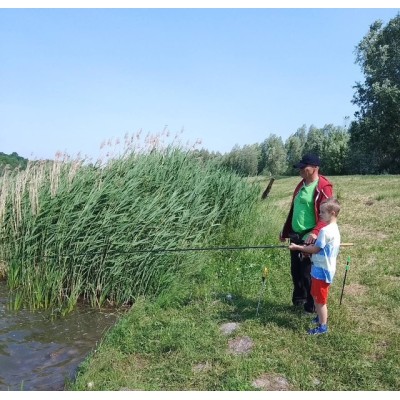 Mężczyzna z dzieckiem wędkują nad brzegiem zbiornika wodnego otoczonego trawą i roślinnością.
