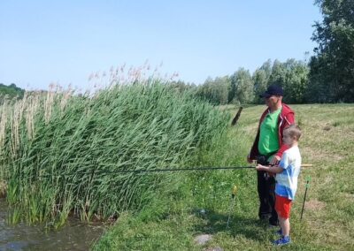 Mężczyzna z dzieckiem wędkują nad brzegiem zbiornika wodnego otoczonego trawą i roślinnością.