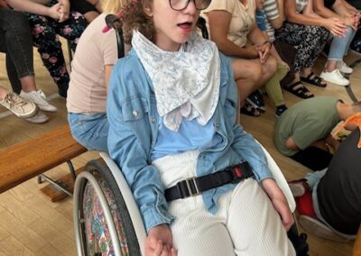 Osoba na wózku inwalidzkim z kapeluszem w kształcie ryby siedzi na sali gimnastycznej.