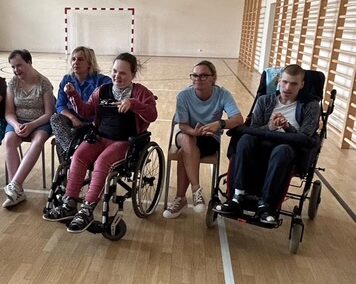 Osoby, w tym dwie na wózkach, siedzą na krzesłach w hali sportowej z boiskiem.