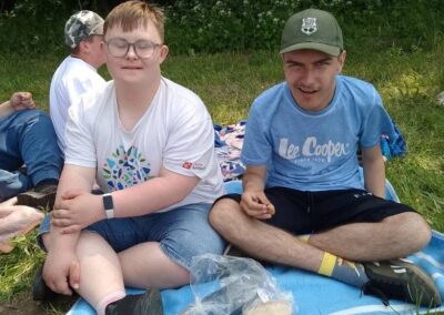 Dwie osoby siedzące na kocu na trawie, przed nimi przezroczysta torba z pieczywem i butelka.