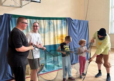 Pięć osób w sali gimnastycznej trzyma dużą siatkę rybacką na tle zasłony morskiej i tablicy do koszykówki.