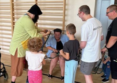 Grupa osób pomaga kobiecie wyjść z siatki w sali gimnastycznej z drabinkami na ścianie.