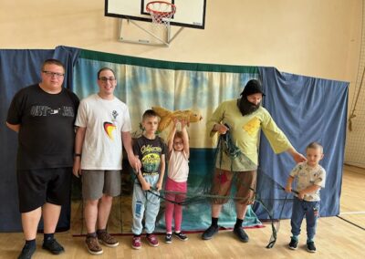 Grupa sześciu osób w sali gimnastycznej z kostiumem rybaka, maskotką ryby i siatką.