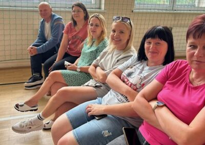 Sześć osób siedzi na ławkach w sali sportowej obok dużych okien z siatką na ścianie.