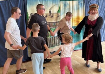 Osoby trzymające się za ręce i tańczące w kole na sali gimnastycznej z dekoracją wiejskiej chaty.