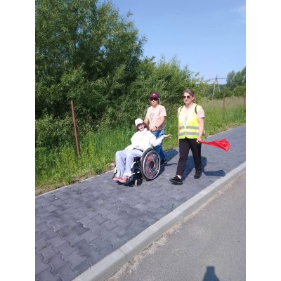 Trzy kobiety idą chodnikiem, jedna na wózku, druga prowadzi wózek, trzecia w kamizelce trzyma czerwoną chorągiewkę.