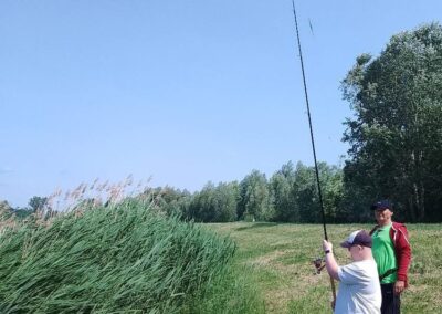 Dwaj mężczyźni nad wodą, jeden trzyma wędkę, otoczeni zielenią i drzewami.