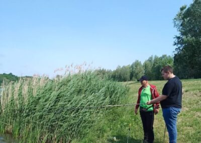 Dwóch mężczyzn łowi ryby nad zbiornikiem wodnym otoczonym trawą i trzcinami przy słonecznej pogodzie.