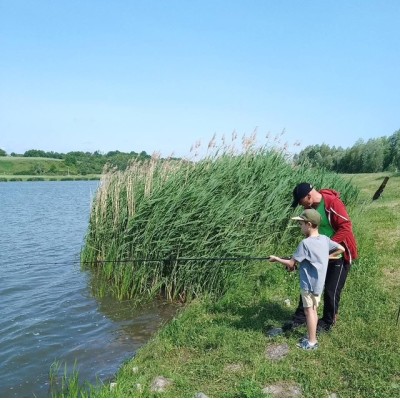 Dwoje osób wędkuje nad brzegiem jeziora otoczonego roślinnością, przy słonecznej pogodzie.