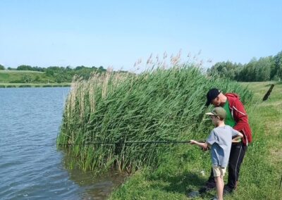 Dwoje osób wędkuje nad brzegiem jeziora otoczonego roślinnością, przy słonecznej pogodzie.