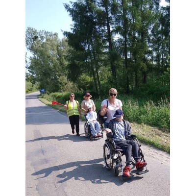 Dwie osoby na wózkach inwalidzkich i dwie osoby ich wspierające idą drogą przy lesie.