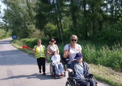 Dwie osoby na wózkach inwalidzkich i dwie osoby ich wspierające idą drogą przy lesie.