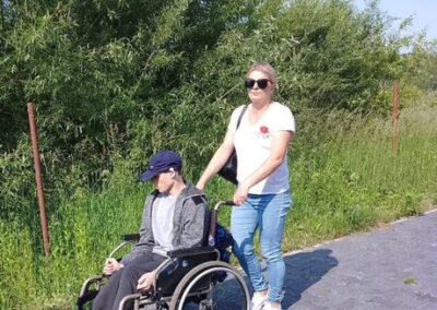 Kobieta pcha mężczyznę na wózku inwalidzkim po chodniku obok zieleni.