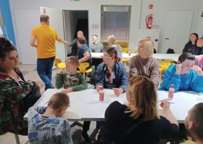 Dzieci i dorośli siedzą przy białych stołach w jasnym pokoju z żółtymi krzesłami.