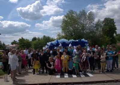Grupa dzieci i dorosłych na drewnianej platformie z dekoracją z niebiesko-białych balonów na zewnątrz.