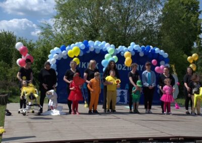 Dzieci i dorośli na drewnianej scenie z kolorowymi balonami i dekoracjami na zewnątrz.