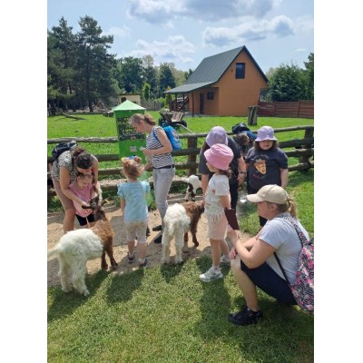 Grupa dzieci i dorosłych w zagrodzie z młodymi kozami przy domu i ogrodzeniu drewnianym.