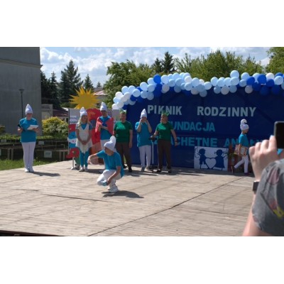 Osoby w kostiumach smerfów na drewnianej scenie podczas rodzinnego pikniku z niebieską dekoracją.