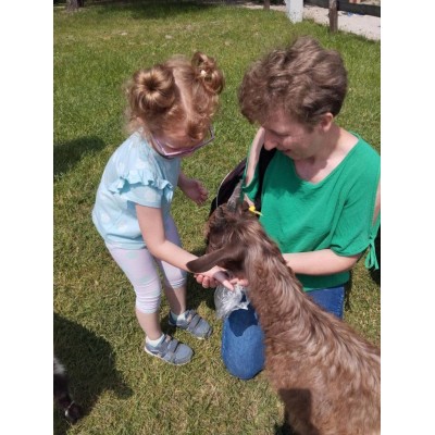 Dziewczynka i kobieta karmią kozę na trawniku w słoneczny dzień.