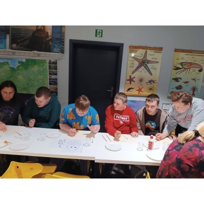 Grupa osób maluje przy stole z przyborami, w tle edukacyjne plakaty morskich organizmów.