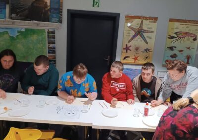 Grupa osób maluje przy stole z przyborami, w tle edukacyjne plakaty morskich organizmów.