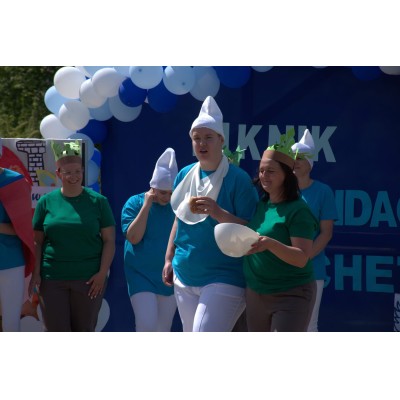 Grupa osób w kostiumach, niebieskie i białe balony w tle, jedna trzyma pączka, inna miskę.