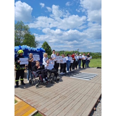 Grupa ludzi na drewnianej platformie z kartkami, w tym strażak i osoby na wózkach inwalidzkich.