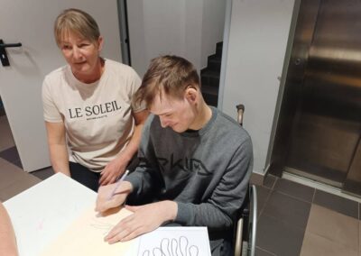 Mężczyzna na wózku inwalidzkim rysujący przy stole, obok kobieta obserwująca, w tle winda i schody.