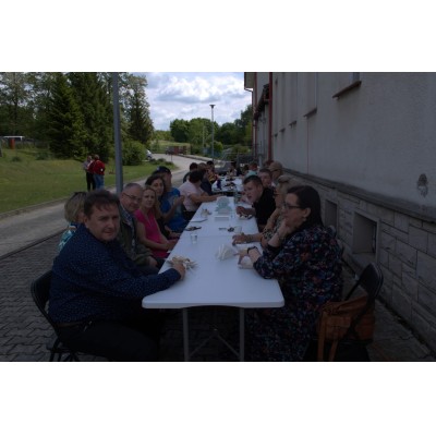 Ludzie siedzący przy długim stole na zewnątrz budynku, jedzący i rozmawiający.