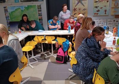 Osoby przy stołach w sali edukacyjnej malują obrazy, na ścianach plansze i mapa.