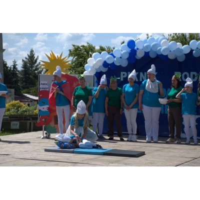 Grupa osób w kostiumach Smerfów na scenie, jedna osoba klęczy przy leżącym dziecku.