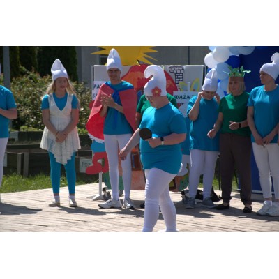 Grupa osób w kostiumach smerfów na drewnianej scenie na zewnątrz podczas wydarzenia.