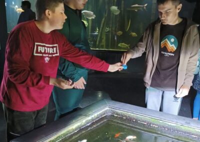 Trzech mężczyzn karmi ryby w akwarium z różnorodnymi gatunkami ryb w ciemnym pomieszczeniu.