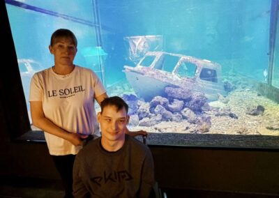 Kobieta i mężczyzna na wózku inwalidzkim na tle akwarium z zatopioną łodzią i dekoracjami.