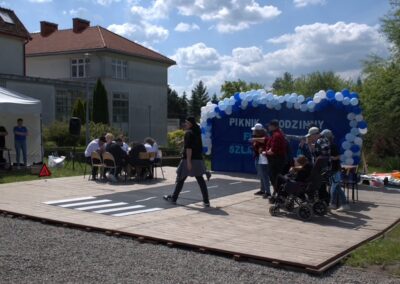 Drewniana platforma z makietą przejścia, grupą ludzi, balonami i banerem na pikniku.