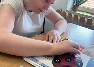 Uczestnik przy stole układa i przykleja papierowe kółka z literami na ilustracji biedronki na karcie pracy.